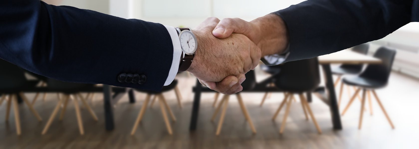 Two people in business suits shaking hands across a meeting room table